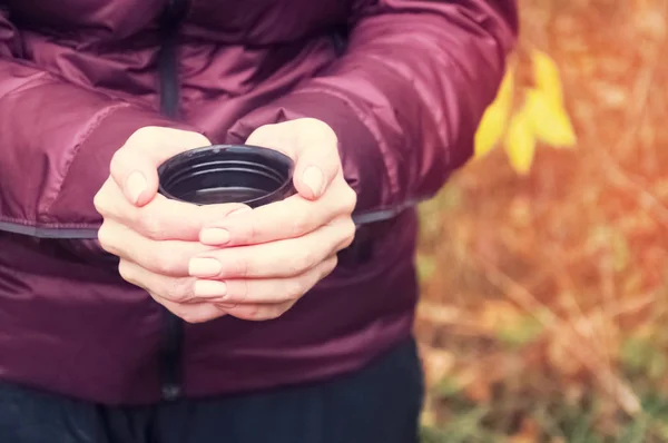 A girl in a lilac jacket holds a glass from a thermos with hot tea. In the autumn park