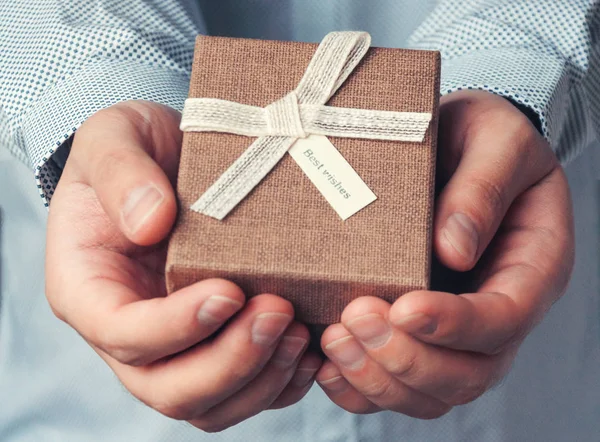 A gift in the hands of man. Close-up a brown gift box and a white ribbon with a tag. Toning and blur