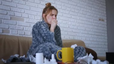 Güzel beyaz kadın hasta, kanepede oturuyor. Burnu tıkalı, hapşırıyor ve öksürüyor. Burnunu sümkür ve gri örtülerin altına saklan..