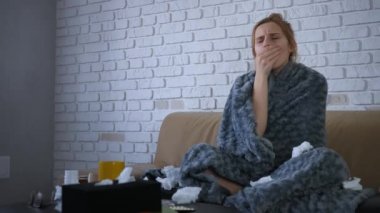 Güzel beyaz kadın hasta, kanepede oturuyor. Burnu tıkalı, hapşırıyor ve öksürüyor. Burnunu sümkür ve gri örtülerin altına saklan..
