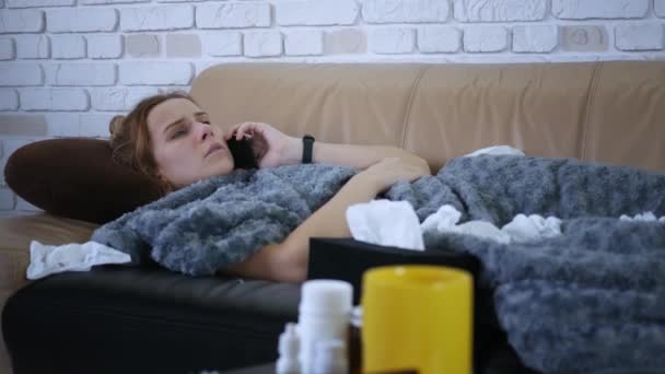 病気の若い白人女性は 医師とオンラインで話しています毛布の下にソファの上に横たわるスマートフォンに錠剤を示すビデオ協議を得ています 人と道具の概念 — ストック動画
