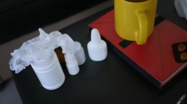 Kâğıt peçete alan beyaz bir kadının elleri. Bir fincan çay, hap, kitap, termometre, kağıt havlu dolu bir masa. İçeride. Tedavi için. Hastalık. Kapat..