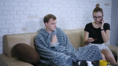 Kanepede oturan genç bir çift. Kadın soğuk algınlığı için ilaç tutuyor, aile doktorunu arayıp hasta bir adama bakmakla ilgili tavsiye istiyor..