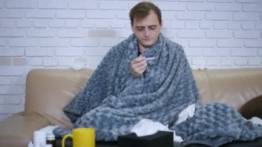 Genç beyaz adam kanepede otururken kendini kötü hissediyor ve sonra vücut ısısını termometre kullanarak kontrol ediyor. Hastalıklı kişi hastalığı virüs portresi