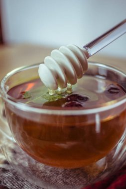 Silikon ballı ve balı cam çay bardağına daldıran kuru gül kalçası ve ahşap masa üzerinde taze nane yaprağı renkli arka plan, bitkisel çay ve sağlıklı içecek konsepti. Kapat..