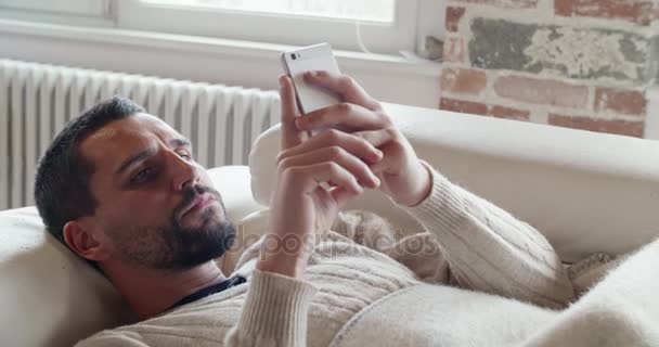 Jovem caucasiano homem detalhe relaxante no sofá usando smartphone indoor na moderna casa industrial. 4k handheld close up vídeo tiro — Vídeo de Stock