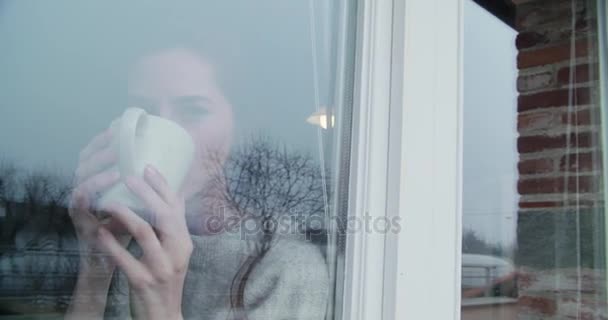 Jonge Kaukasische vrouw drinken kopje koffie of thee, ontspannen op zoek buiten venster binnen in industriële stadsvilla. 4 k handbediende buiten reflecties bekijken video schot — Stockvideo