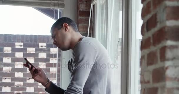 Joven hombre negro relajante con teléfono inteligente y mirando fuera de la ventana interior en la casa industrial moderna. 4k handheld close up video shot — Vídeo de stock