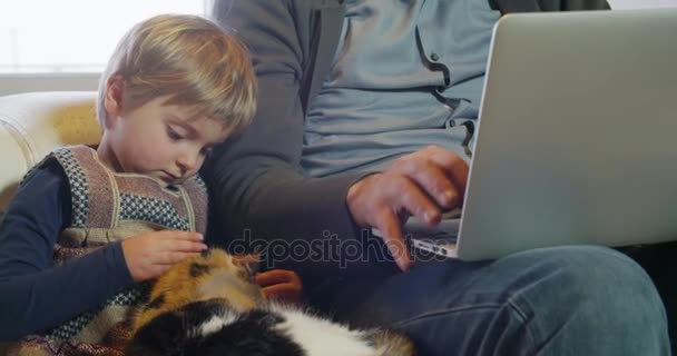 Família moderna menina brincando com gato, enquanto o pai trabalha com notebook no sofá interior na casa industrial moderna. caucasiano. 4k handheld close up câmera lenta de vídeo — Vídeo de Stock