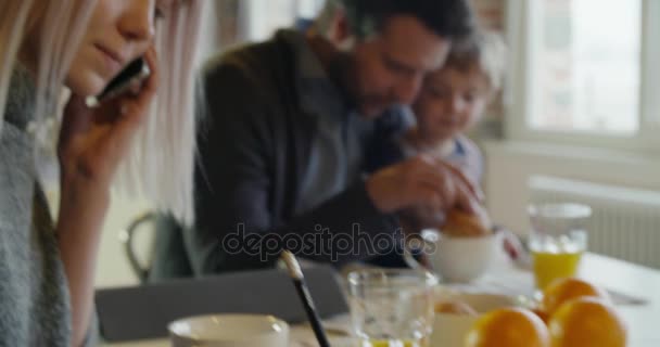 Modernes Familienmädchen frühstückt mit Papa, während Mama das Smartphone nutzt und drinnen im modernen Industriehaus arbeitet. Kaukasisch. 4k Handheld-Zeitlupenvideo gedreht — Stockvideo