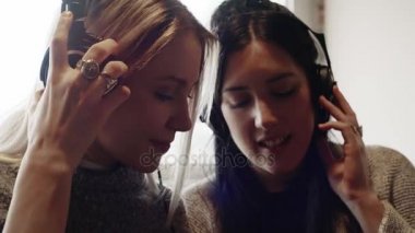 Genç Beyaz sarışın kadın ve arkadaşı modern endüstriyel evde kanepede kapalı kulaklık ile müzik dinlemek. 4 k el video çekim