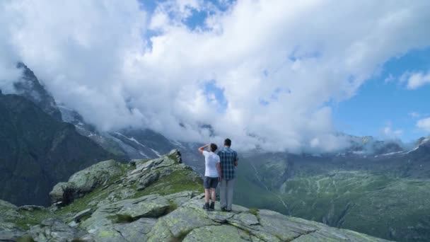 Wanderlust moviéndose hacia atrás sobre dos personas que miran el valle de la montaña en verano soleado con nubes. Europa Alpes naturaleza verde al aire libre paisaje montañas vuelo aéreo salvaje establishment. — Vídeos de Stock