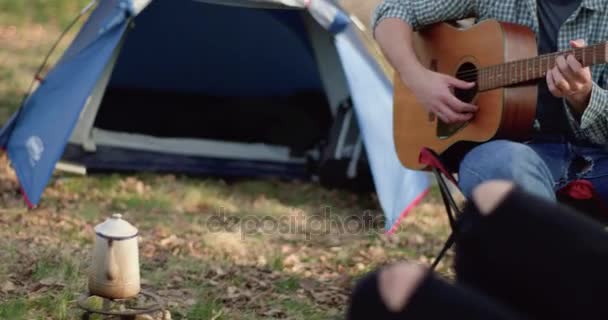 Hombre relajante tocando la guitarra cerca de la chimenea con amigos beverage.real caliente gente al aire libre camping tienda de campaña vacaciones en viaje de otoño.Otoño día soleado en la naturaleza, la unión y la amistad. — Vídeos de Stock