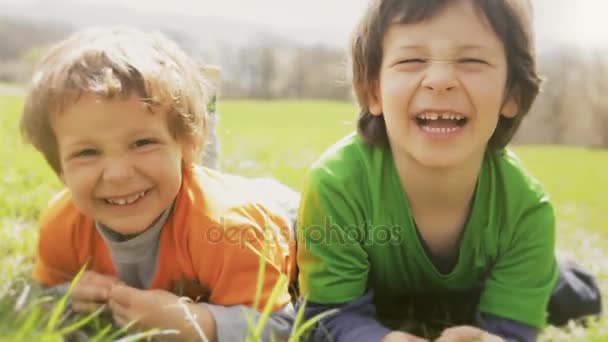 Niños felices sonriendo de cerca retrato. Dos hermanos niños riendo y jugando mientras se acuestan en el prado de hierba en el día soleado al aire libre. Familia hijo chicos relajarse en la naturaleza. material de vídeo — Vídeo de stock