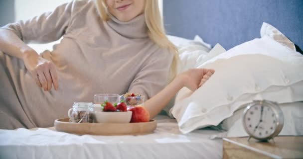 Schöne Frau beim Frühstück mit Joghurt, Obst und Müsli auf dem Bett. Morgen aufwachen zu Hause im Schlafzimmer. Kaukasier, die auf dem Bett liegen. 4k-Video — Stockvideo