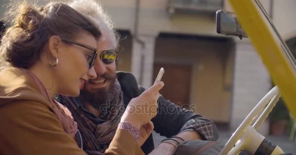 Γυναίκα με γυαλιά ηλίου χαμογελώντας χρησιμοποιώντας με έξυπνο κινητό τηλέφωνο με το φίλο της. Πλευρά βολή, ήλιος οπίσθιου φωτισμού. Καυκάσιος ζευγάρι στην αγάπη roadtrip παραθεριστικές κατοικίες ιταλική ταξίδια σε μετατρέψιμο vintage car.4k χειρός βίντεο — Αρχείο Βίντεο