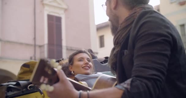 Man smiling while playing guitar music with girlfriend.Woman listening to boyfriend singing song.Caucasian couple in love roadtrip vacation italian travel on convertible vintage car.4k handheld video — Stock Video