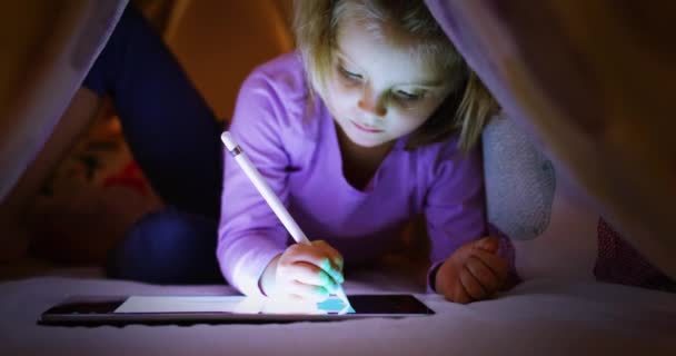 Детская блондинка-кавказка цифровой раскраски с планшетным приложением под одеялом ночью. Современные технологии дети используют дома. 4k видео — стоковое видео