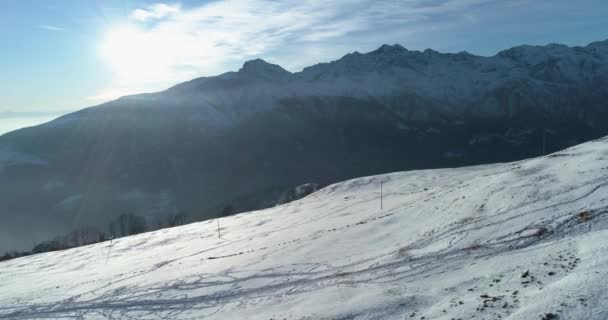 Achterwaartse luchtfoto bovenaanzicht over winter besneeuwde berg met ski tracks in zonnige dag met wolken. Witte achtergrondverlichting Alpen bergen sneeuw seizoen oprichter met zon shine.4k drone vlucht tot oprichting van schot — Stockvideo