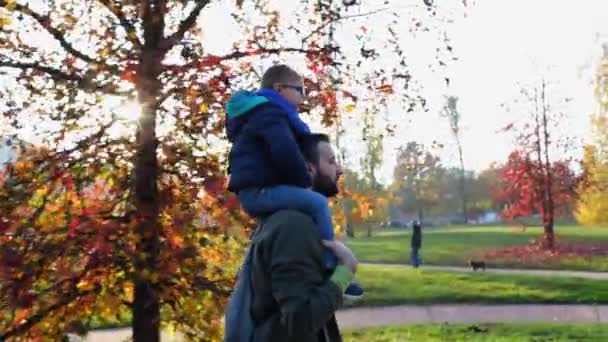 Vader lopen in stedelijke stadspark met zoon op de schouders. Vader een kind genieten van een buiten herfstdag samen. Zonsondergang, verlicht. Saamhorigheid, familie en kindertijd concept.4k kant Volg video schot — Stockvideo