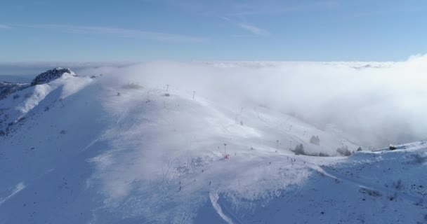 Voorwaartse antenne over winter besneeuwde berg top ski tracks resort met skiër mensen skiën. Zonnige dag, mistige wolken. Mist stijgt. Bergen van de Alpen sneeuw seizoen establisher.4k drone vlucht sport tot oprichting van — Stockvideo