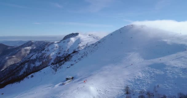Κεραία προς τα πίσω πάνω από χειμώνα χιονισμένο βουνό Κορυφαία χιονοδρομικά κομμάτια θέρετρο με ανθρώπους σκιέρ σκι. Ηλιόλουστη μέρα, ομιχλώδης σύννεφα. Ομίχλη αυξάνεται. Άλπεις Όρη χιόνι σεζόν establisher.4k κηφήνας πτήσης άθλημα για την ίδρυση — Αρχείο Βίντεο
