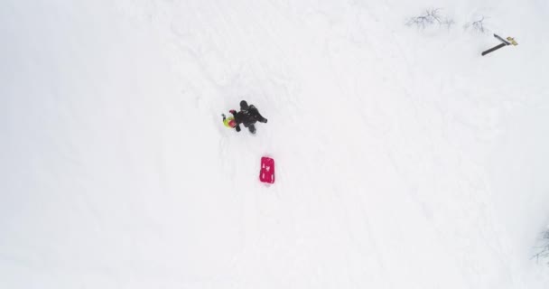 Зимовий повітряний вид зверху над батьком тягне червоний бобслей на сніговий підйом з дитиною.Тато, син або дочка, бобслей на снігу. — стокове відео