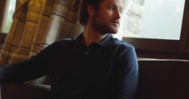 Evdeki oturma odasındaki kanepede dinlenen genç adam. Adam kanepede rahatça oturuyor. Adam eve rahatça bakıyor.