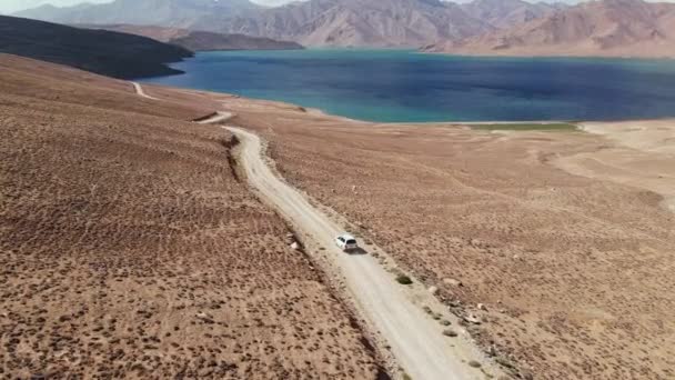 Aerial over road 4x4 car driving along gravel trail path towards lake in arid mountains.Pamir Autópálya selyemút utazó kaland Tádzsikisztánban, Ázsiában.Khorugh Bulunkul tó. 4k drónrepülős videó — Stock videók