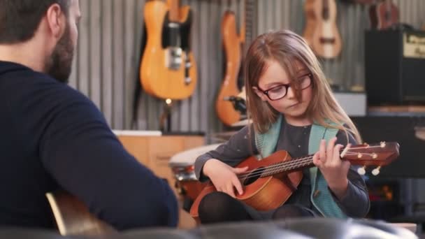 Baba kızına gitar ve ukulele öğretiyor. Küçük kız evde gitar öğreniyor. Ukulele dersini evde kapat. Çocuk babasından gitar öğreniyor. — Stok video