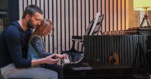 Papà insegna pianoforte alla figlia.Bambina che impara il pianoforte a casa.Vista laterale.Lezione di pianoforte a casa. Bambino che impara il pianoforte da suo padre — Foto Stock