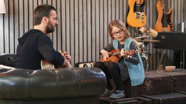Dad teaching guitar and ukulele to his daughter.Little girl learning guitar at home.Side view.Ukulele class at home. Child learning guitar from her father