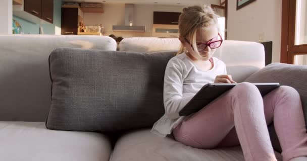 Kind mit Tablet zu Hause auf der Couch sitzend. Kleines Mädchen, das auf dem Sofa sitzt und Hausaufgaben mit Tisch.Frontansicht macht. Kind spielt mit Tablet im Wohnzimmer — Stockvideo