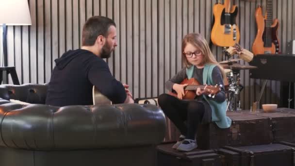 Papa bringt seiner Tochter Gitarre und Ukulele bei. Kleines Mädchen lernt zu Hause Gitarre. Seitenansicht.Ukulele-Kurs zu Hause. Kind lernt Gitarre vom Vater — Stockvideo
