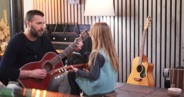 Тато викладає гітару та укулеле своїй дочці. Маленька дівчинка вивчає гітару вдома. Вид збоку. Укулеле клас вдома. Дитяча гітара від батька — стокове відео