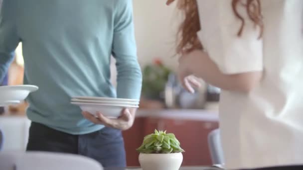 Два человека накрывают стол в гостиной, кладут посуду для еды и разговаривают друг с другом. — стоковое видео