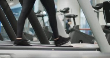 Spor salonunda koşu bandında yürüyen genç bir kadın. Isınmak ya da sakinleşmek. Bacakları yakın çekim, alt vücut detayı. Spor salonunda kadın eğitimi. Kardiyo egzersizi yapan kadın.