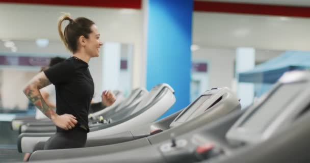Νεαρή γυναίκα που τρέχει σε διάδρομο γυμναστικής. Πλευρική άποψη. Εκπαίδευση γυναικών στο γυμναστήριο. Γυναίκα άσκηση καρδιο προπόνηση — Αρχείο Βίντεο