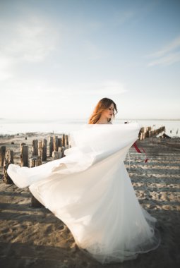 Güzel bayan, gelin gün batımında deniz kenarında bir gelinlik içinde poz veriyor.