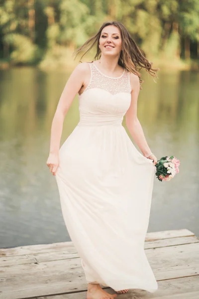 Gyönyörű menyasszony elegáns ruha gazdaság csokor jelentő erdő és tó közelében — Stock Fotó