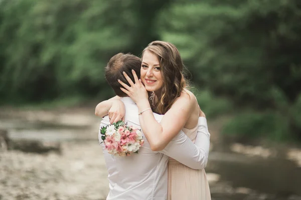 Hermosa pareja de boda besándose y abrazándose cerca de la orilla de un río de montaña con piedras —  Fotos de Stock