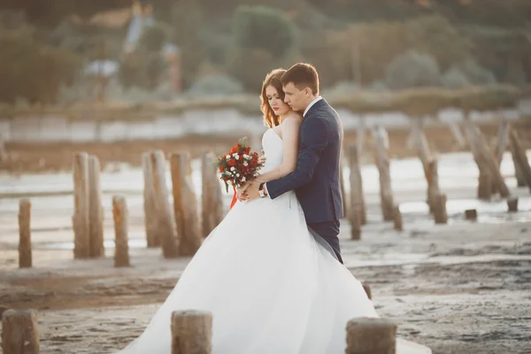 Casal de casamento, noivo, noiva com buquê posando perto do mar ao pôr do sol — Fotografia de Stock