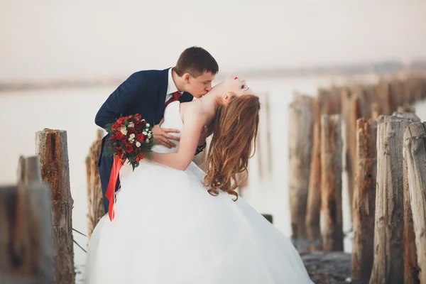 Hermosa joven pareja de boda, novia y novio posando cerca de postes de madera en el mar de fondo —  Fotos de Stock