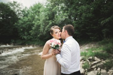 Güzel düğün çifti bir dağ nehrinin kıyısında taşlarla öpüşüp kucaklaşıyorlar.