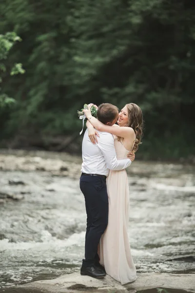 Hermosa pareja de boda besándose y abrazándose cerca de la orilla de un río de montaña con piedras —  Fotos de Stock