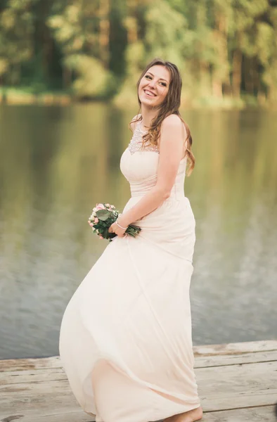 Hermosa novia en vestido elegante celebración de ramo posando cerca del bosque y el lago — Foto de Stock