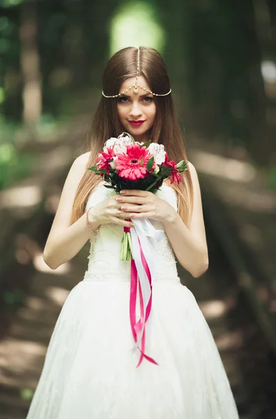 Bella sposa bruna in elegante vestito bianco che tiene bouquet in posa alberi ordinati — Foto Stock