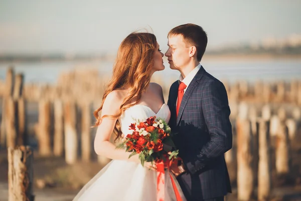 Vackra unga bröllopsparet, bruden och brudgummen poserar nära trästolpar i bakgrunden havet — Stockfoto