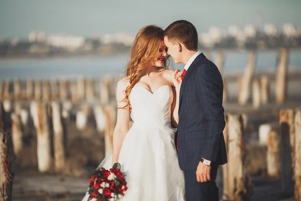 Casal de casamento jovem bonito, noiva e noivo posando perto de postes de madeira no fundo do mar — Fotografia de Stock