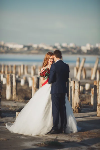 Hermosa joven pareja de boda, novia y novio posando cerca de postes de madera en el mar de fondo — Foto de Stock